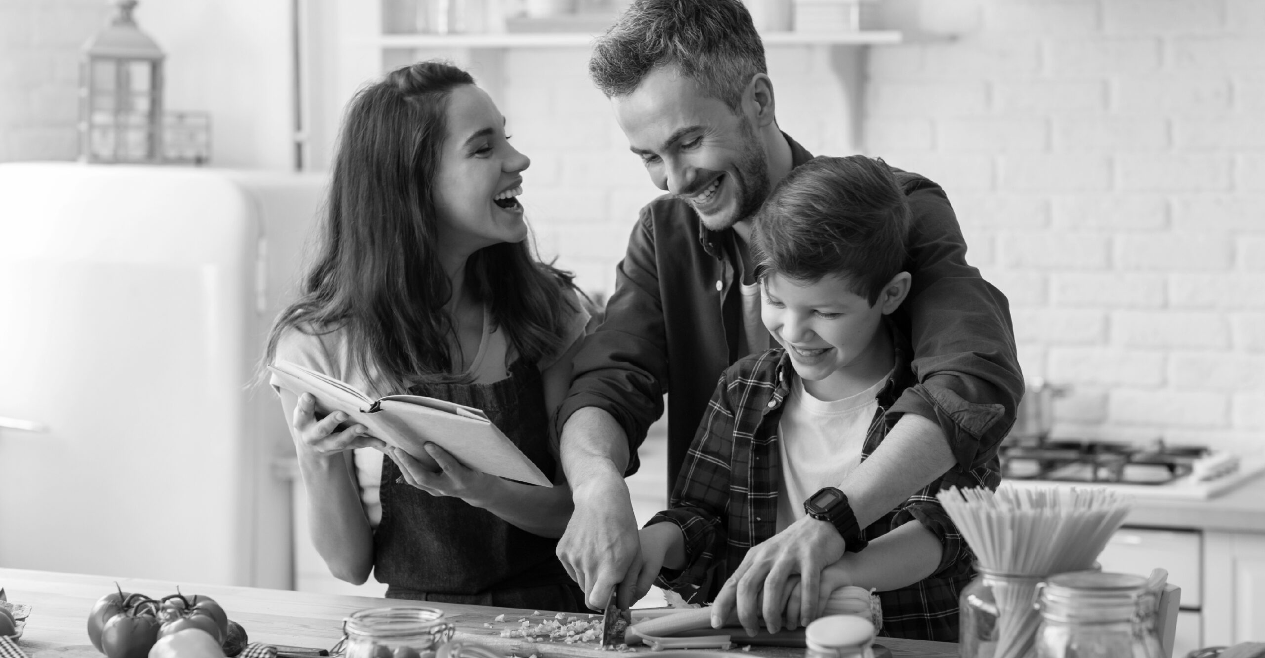 Family of mother, father, and son laughing while the dad helps the son chop food on a cutting board and the mom holds a book